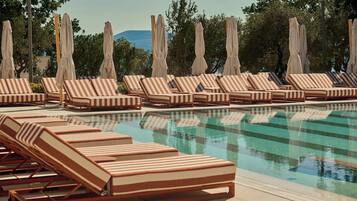 Zona junto a la piscina con sillones reclinables a rayas y sombrillas, rodeada de árboles y un cielo despejado.