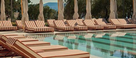 A poolside area with striped lounge chairs and umbrellas, surrounded by trees and a clear sky.