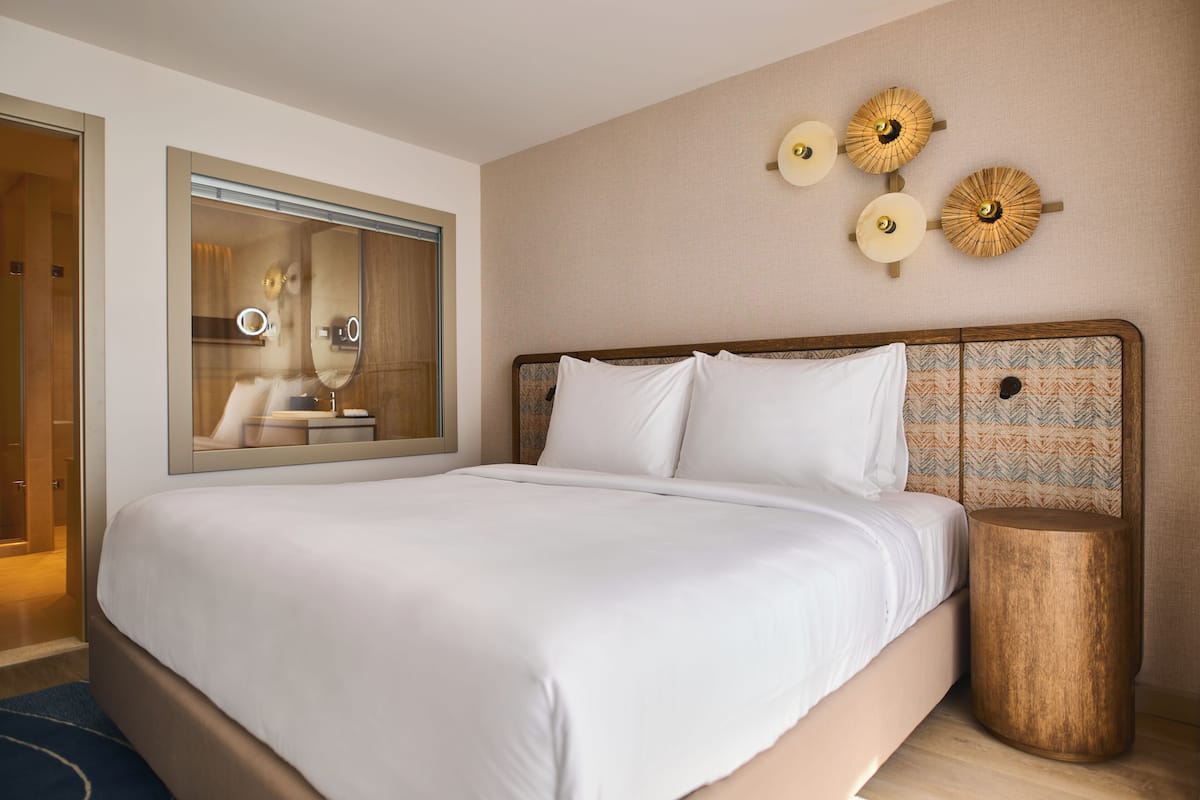 Habitación de hotel con una cama grande, un espejo, una mesita de noche redonda de madera y lámparas fijadas a la pared.