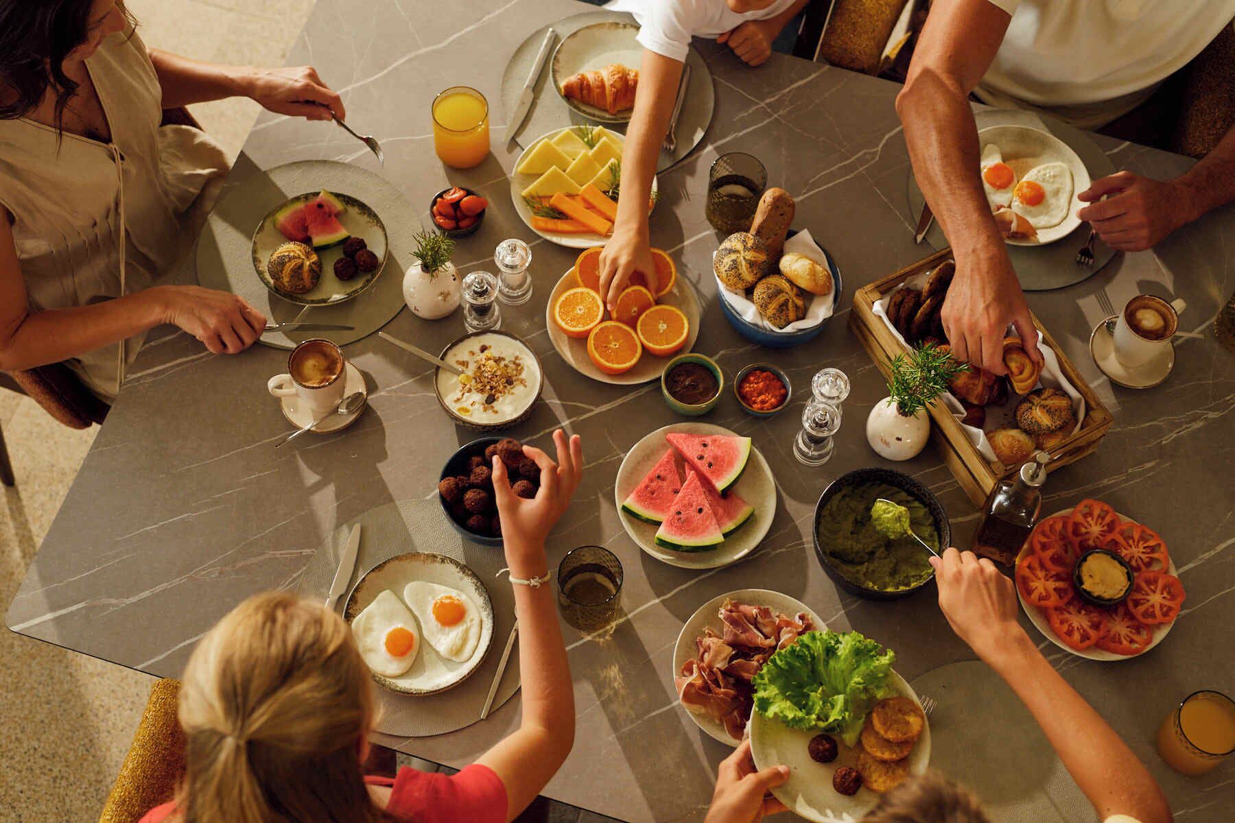 A group of people dining at a marble table with various dishes, including eggs, fruits, and bread.