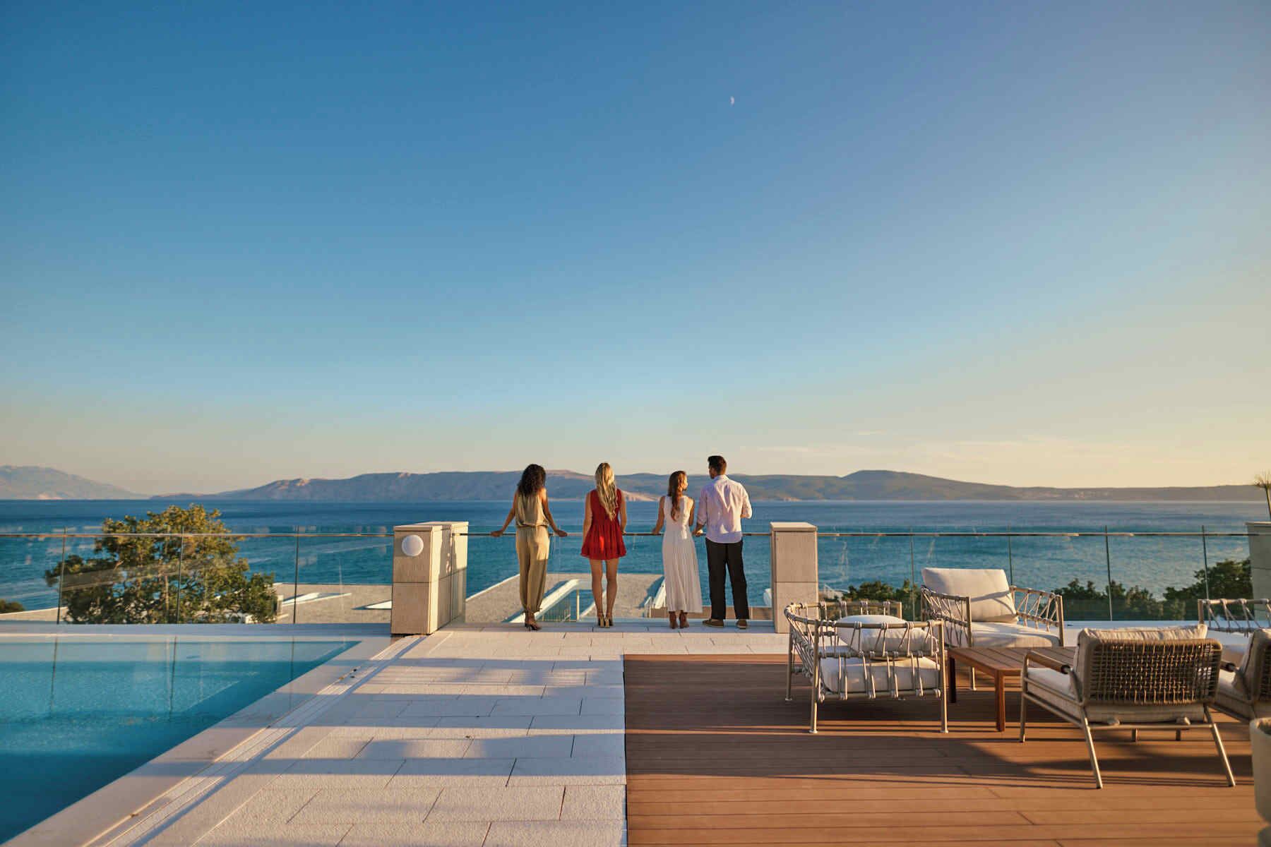 A poolside area with a view of the sea and mountains, outdoor seating, and a clear sky.