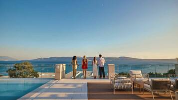 A poolside area with a view of the sea and mountains, outdoor seating, and a clear sky.