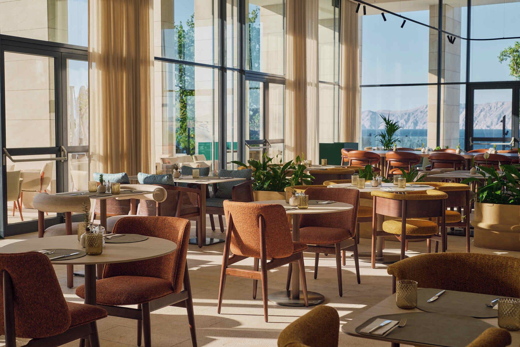 A modern dining area with multiple tables and chairs, large windows with curtains, and indoor plants.