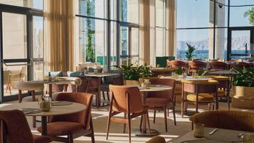 A modern dining area with multiple tables and chairs, large windows with curtains, and indoor plants.
