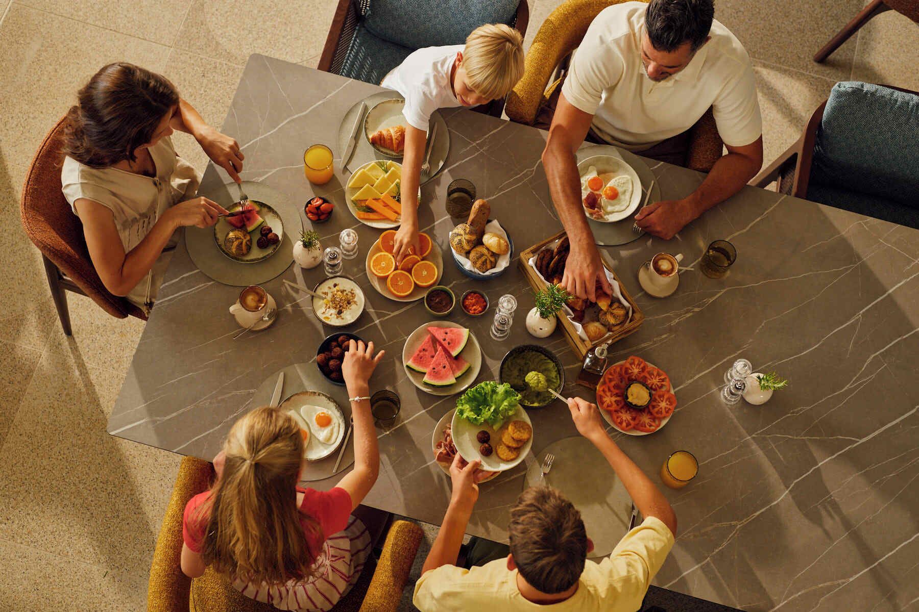 A group of people seated around a table with various dishes, including fruits, bread, and a hot beverage.