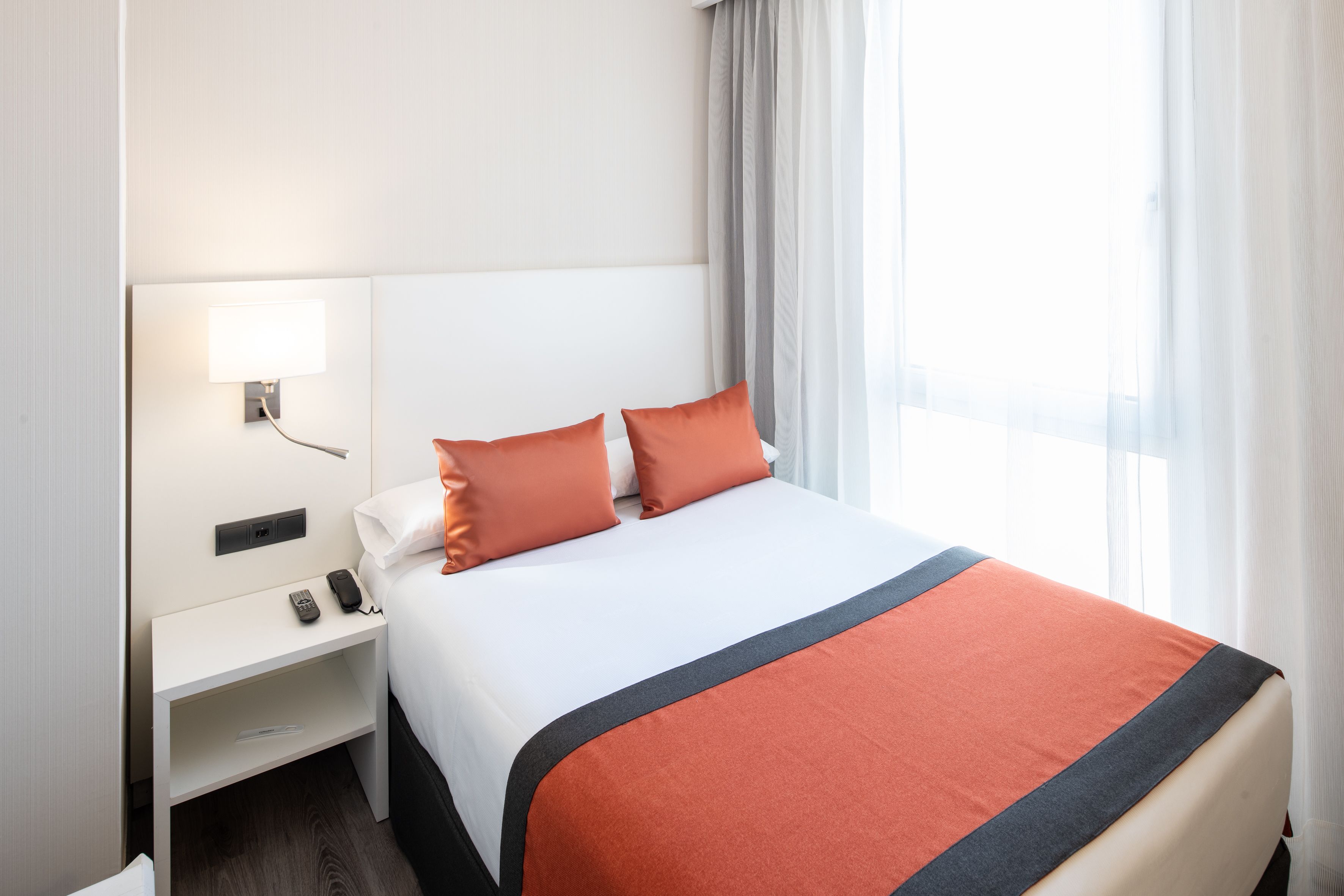 Una habitación de hotel con una cama bien hecha, una mesita de noche con una lámpara y una ventana con cortinas.