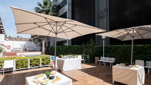 Un espace extérieur aménagé en terrasse, avec des tables disposées pour les repas, de grands parasols offrant de l’ombre et une piscine en arrière-plan.