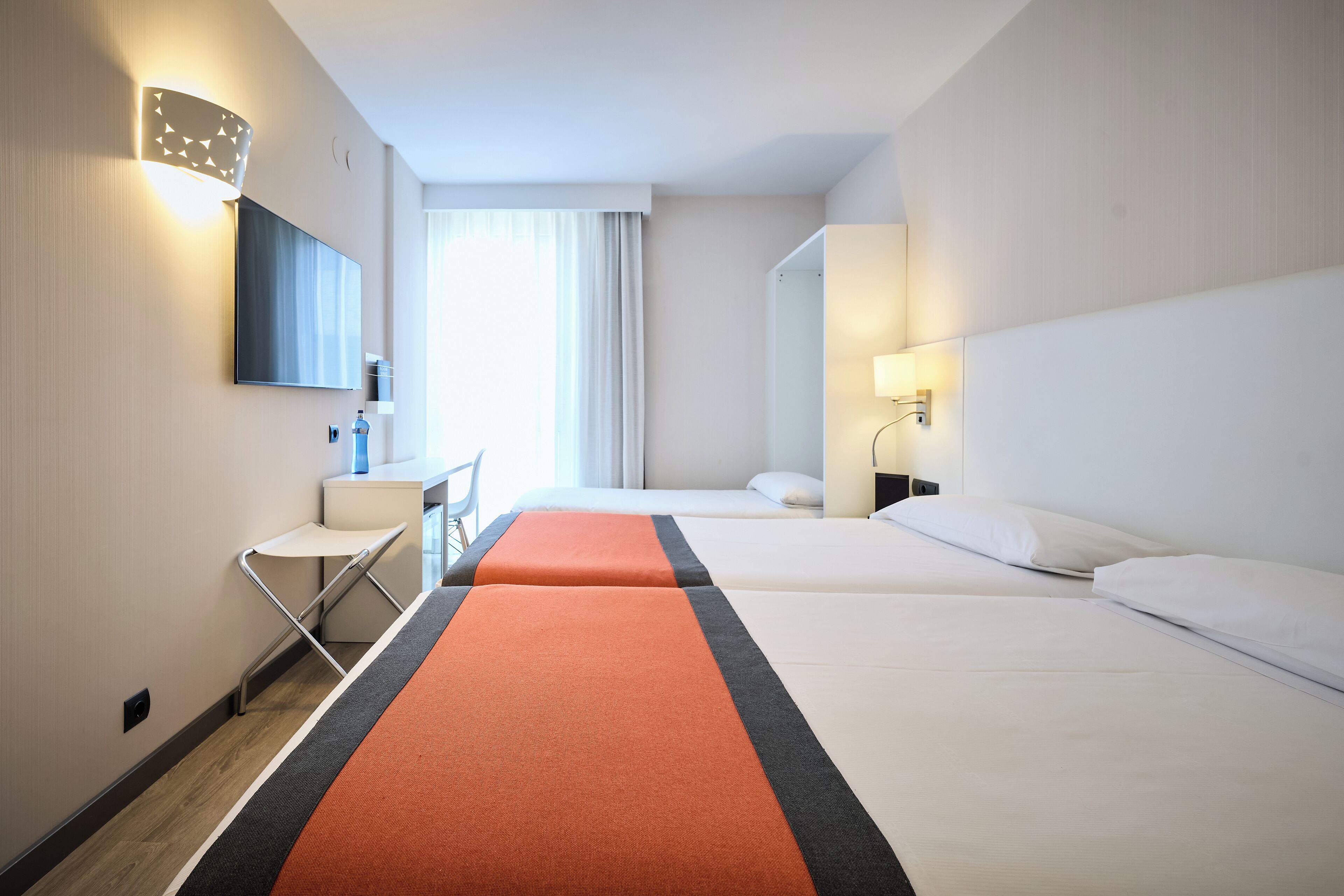 Een hotelkamer met twee bedden, een bureau, een stoel en een groot raam met gordijnen.