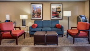 A hotel lobby with a blue sofa, two red armchairs, a brown ottoman, two framed pictures on the wall, a lamp, and an ATM. - Comfort Inn & Suites Lawrenceburg (Lawrenceburg)