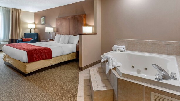 A hotel room with a large bed, a sitting area with a chair, a desk, a desk lamp, a wall-mounted TV, and a separate bathroom with a bathtub. - Comfort Inn & Suites Lawrenceburg (Lawrenceburg)