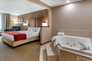 A hotel room with a large bed, a sitting area with a chair, a desk, a desk lamp, a wall-mounted TV, and a separate bathroom with a bathtub.
