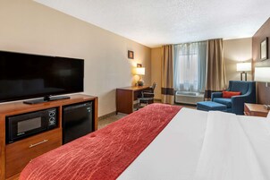 Habitación de hotel con cama, televisor, escritorio con lámpara, silla y ventana con cortinas.