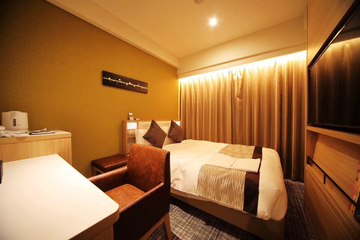 A hotel room with a bed, a desk, a chair, a TV, and a wall-mounted clock.