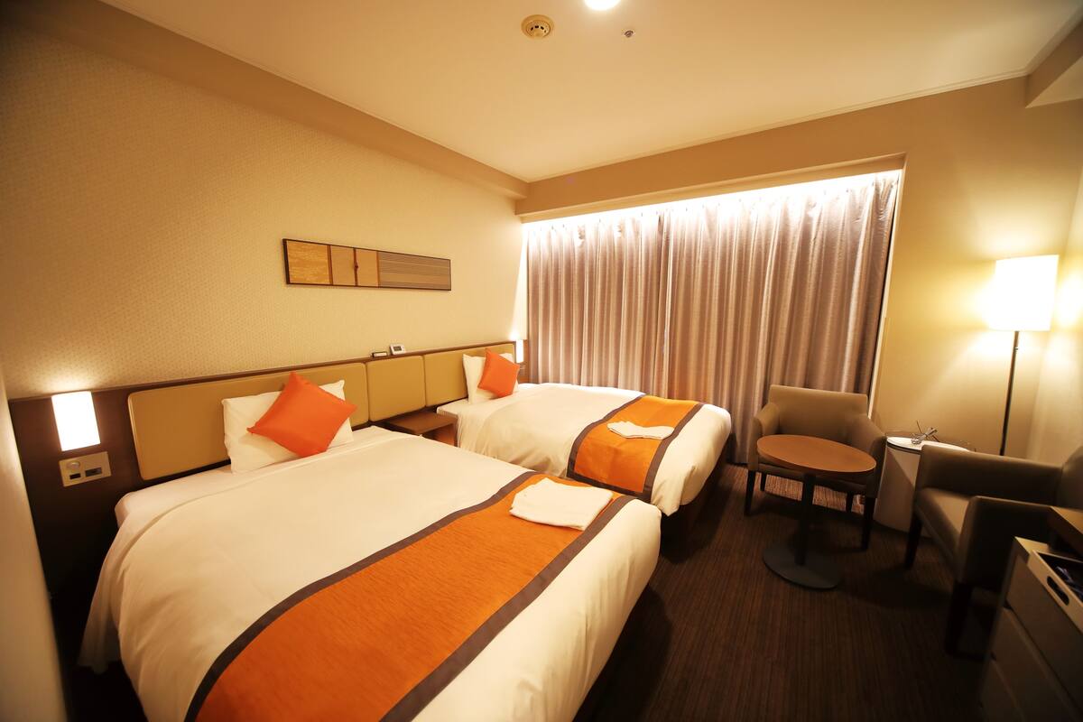 A hotel room with two beds, a small round table, a chair, and a lamp.