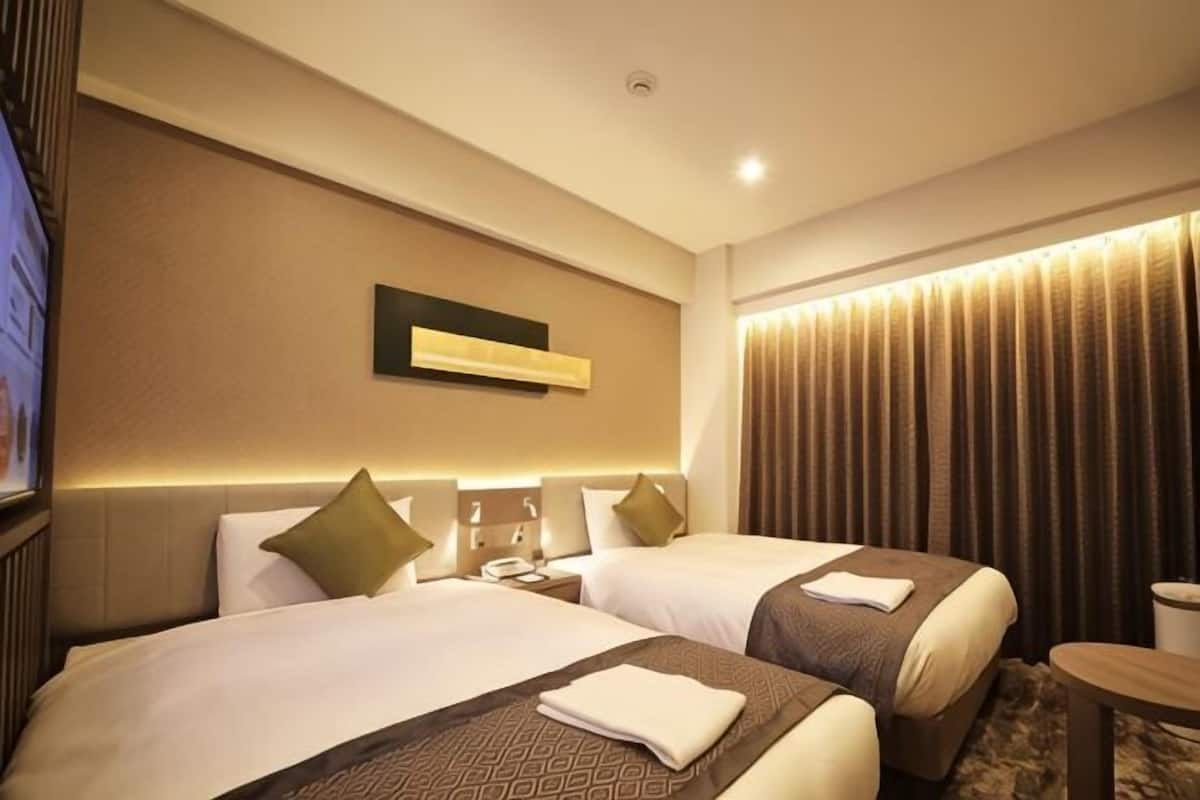 A hotel room with two beds, a flat-screen TV, a bedside table, and a window with curtains.