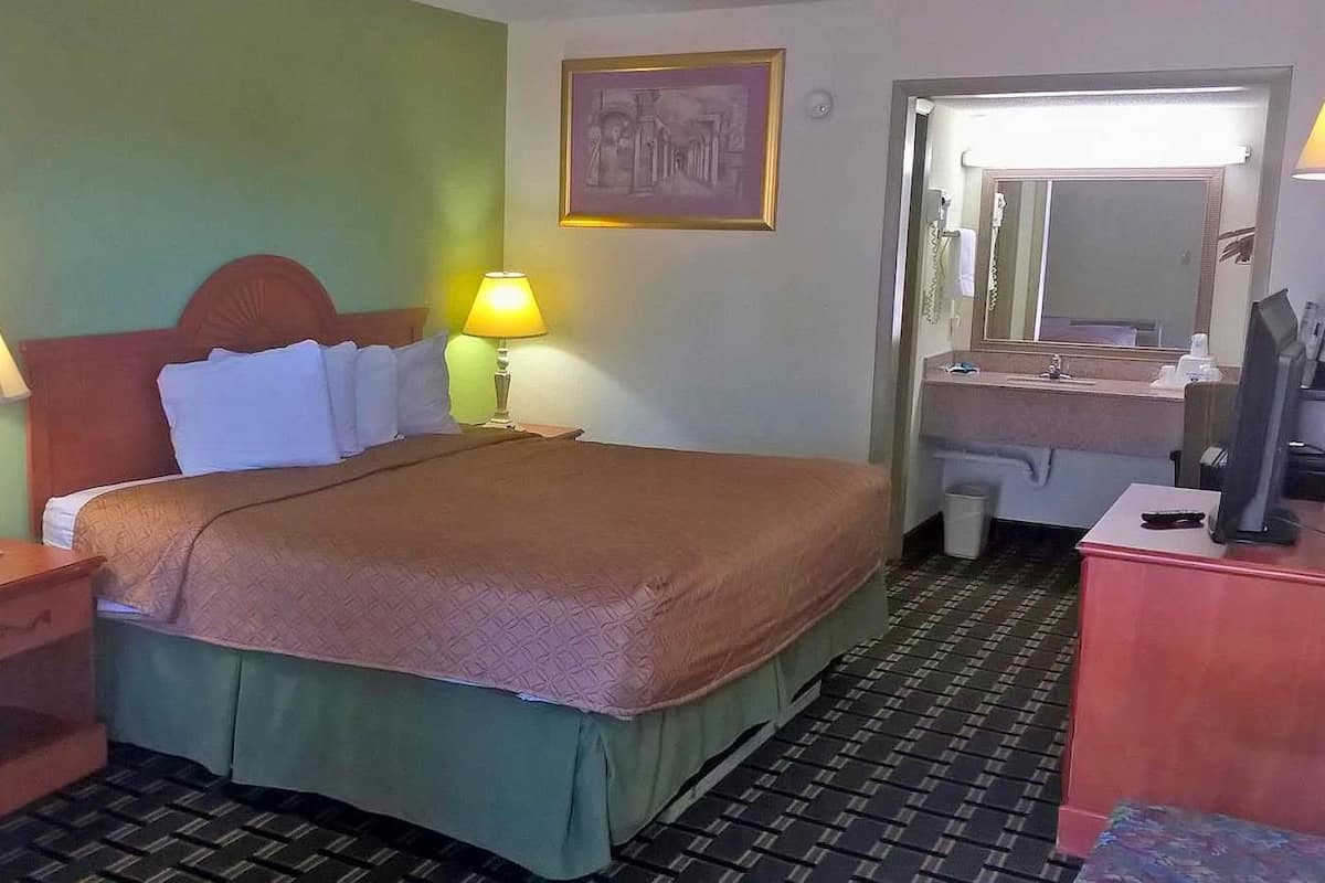A hotel room with a bed, two bedside lamps, a nightstand, a dresser, a mirror, and a bathroom visible through an open door.
