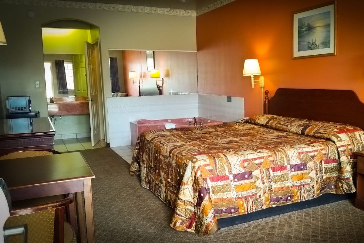 A hotel room with a bed, bedside lamps, a desk, a chair, a bathroom with a mirror, and a painting on the wall.