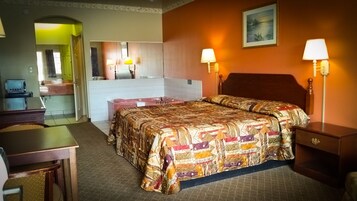 A hotel room with a bed, bedside lamps, a desk, a chair, a bathroom with a mirror, and a painting on the wall.