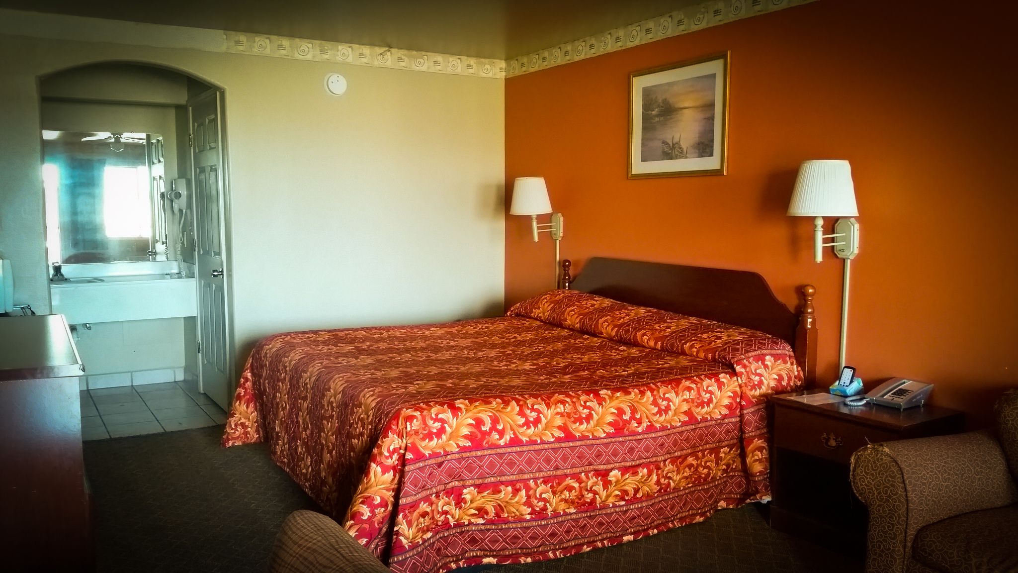 A hotel room with a bed, two bedside lamps, a nightstand, a chair, a painting on the wall, and a bathroom visible through an archway.