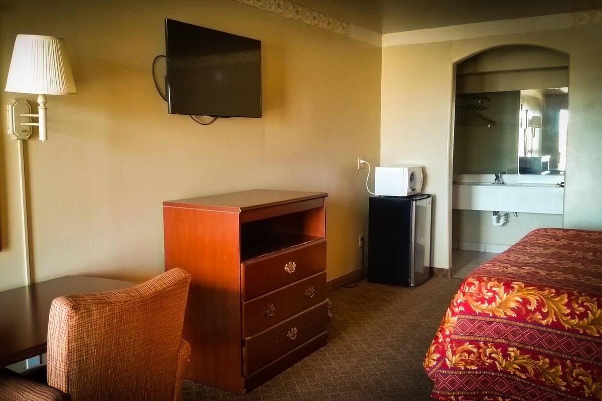A hotel room with a bed, a wooden dresser, a television, a desk with a lamp, and a bathroom visible through an archway.