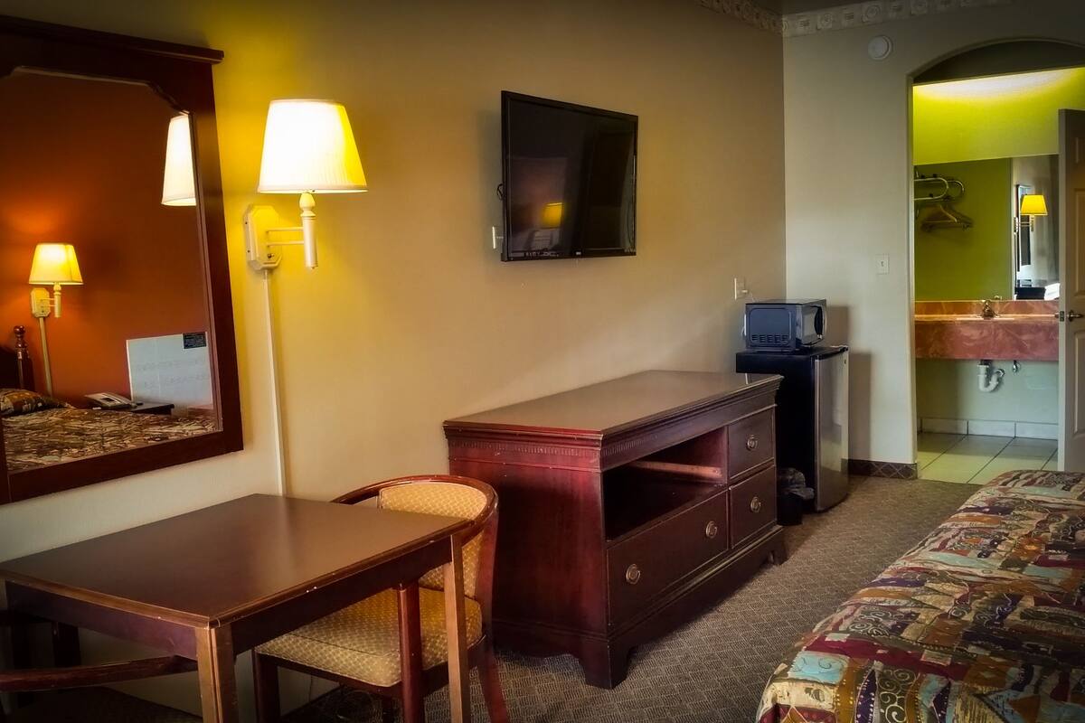 A hotel room with a bed, a desk, a chair, a dresser, a television, and a bathroom visible through an open door.