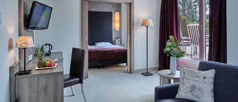 Habitación de hotel con una cama, un escritorio con monitor de computadora, una silla, un sofá, una mesita con lámpara y una ventana con cortinas.