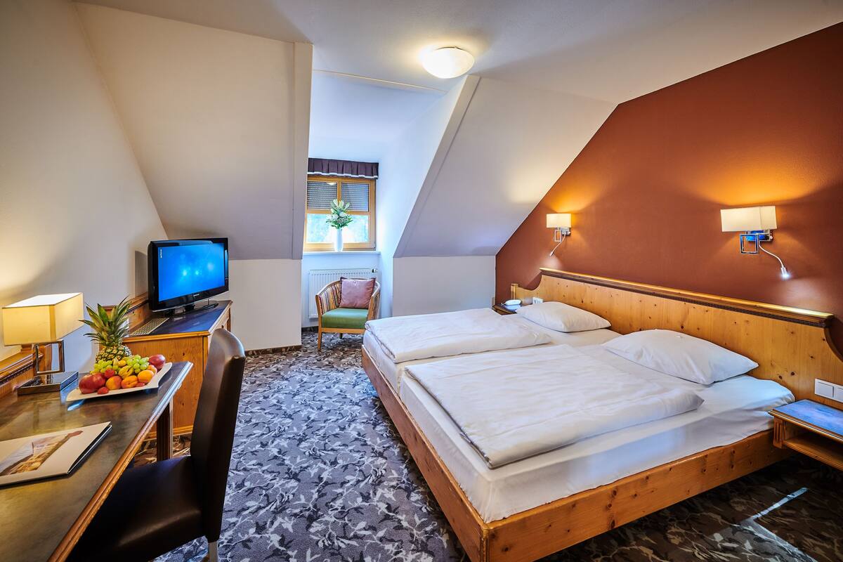 A hotel room with a bed, a desk with a computer, a chair, a television, and a window with a plant.