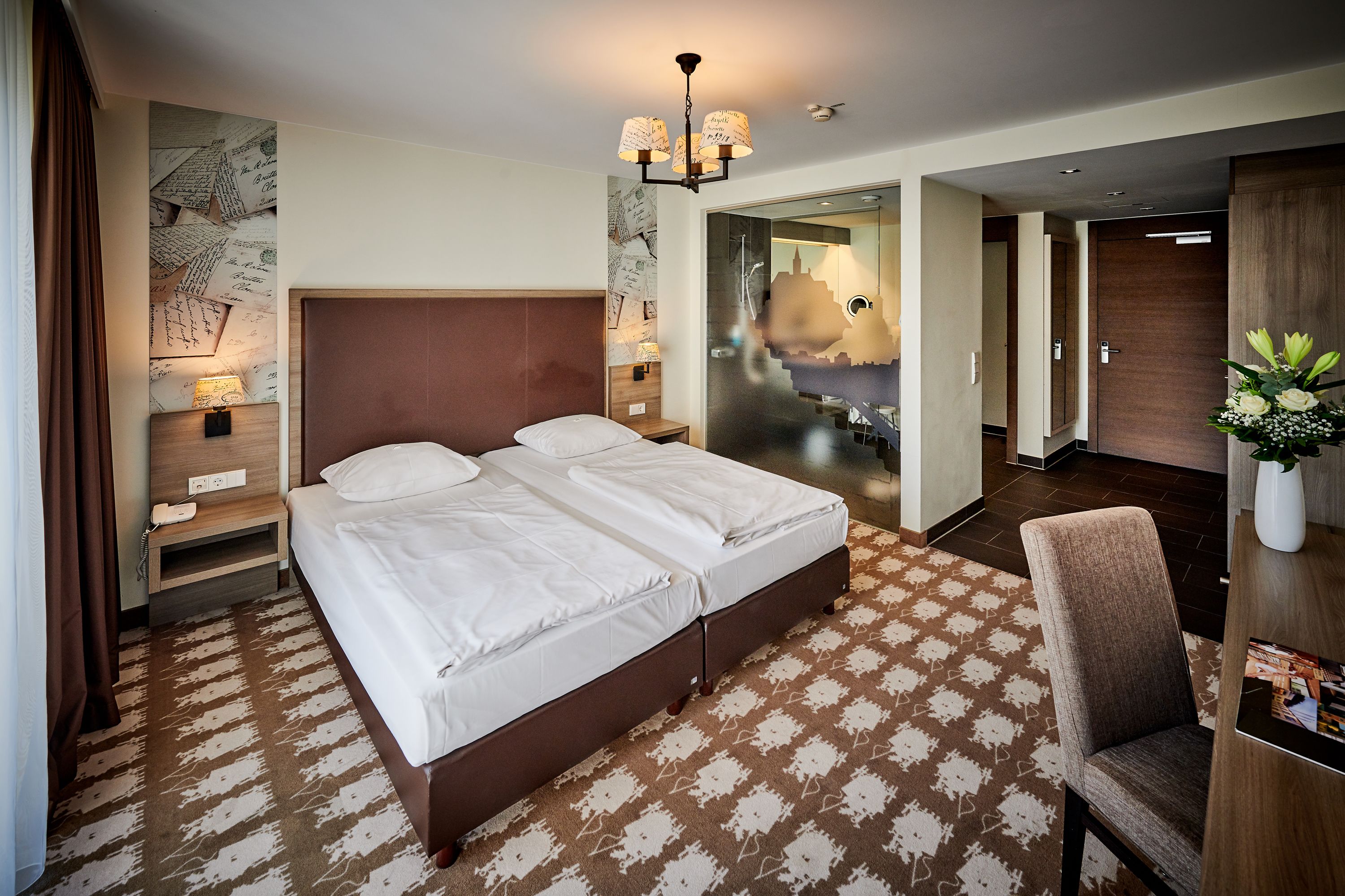 A hotel room with a large bed, a bedside table with a lamp, a chair, and a desk with a vase of flowers.
