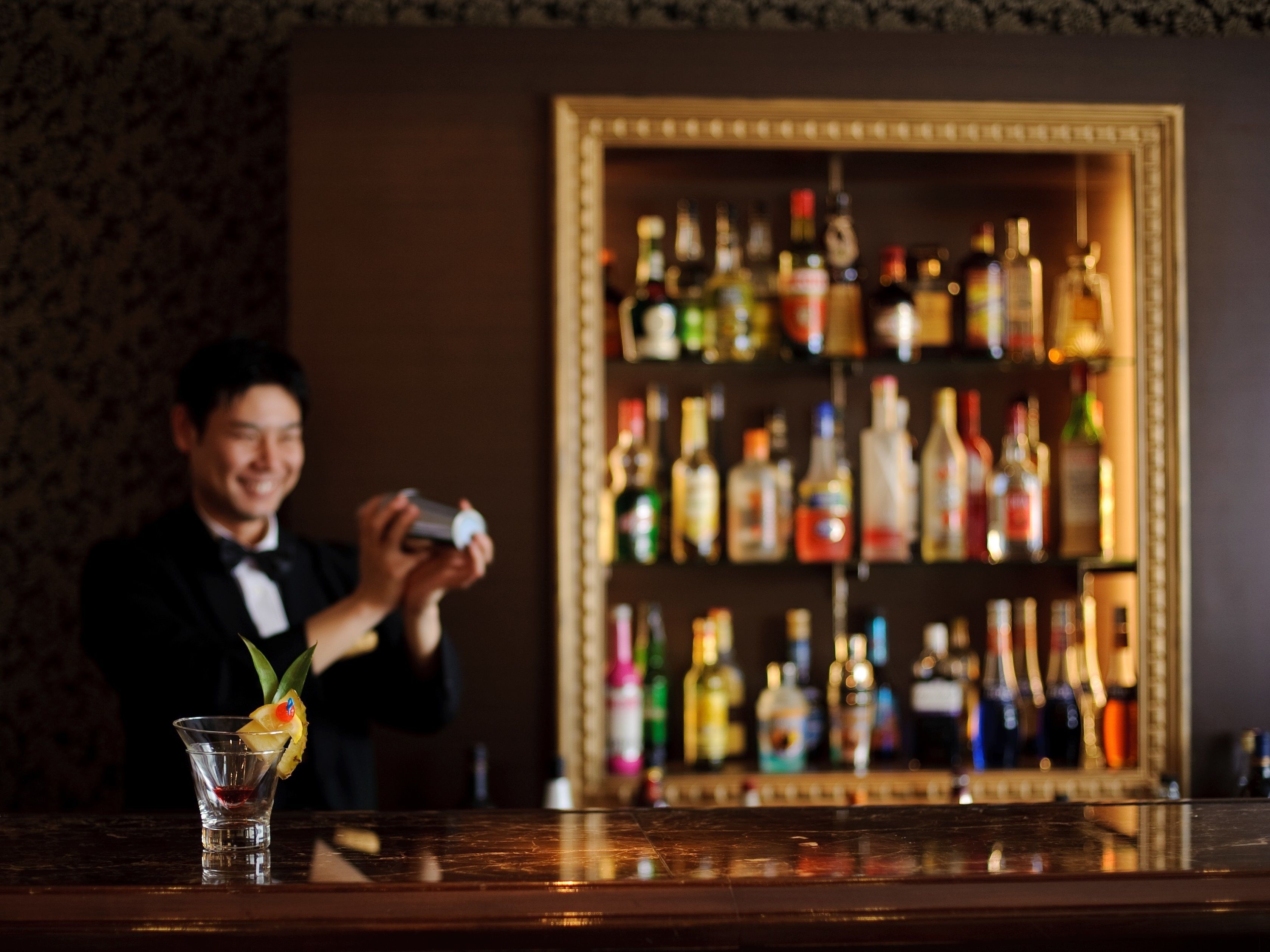 A bar area with a bartender preparing a drink. There is a glass with a garnish on the counter. In the background, there is a shelf with various bottles.