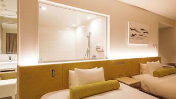 A hotel room with a large window, a bed with white bedding and a green towel, a bedside table with toiletries, and a mirror above the bed.