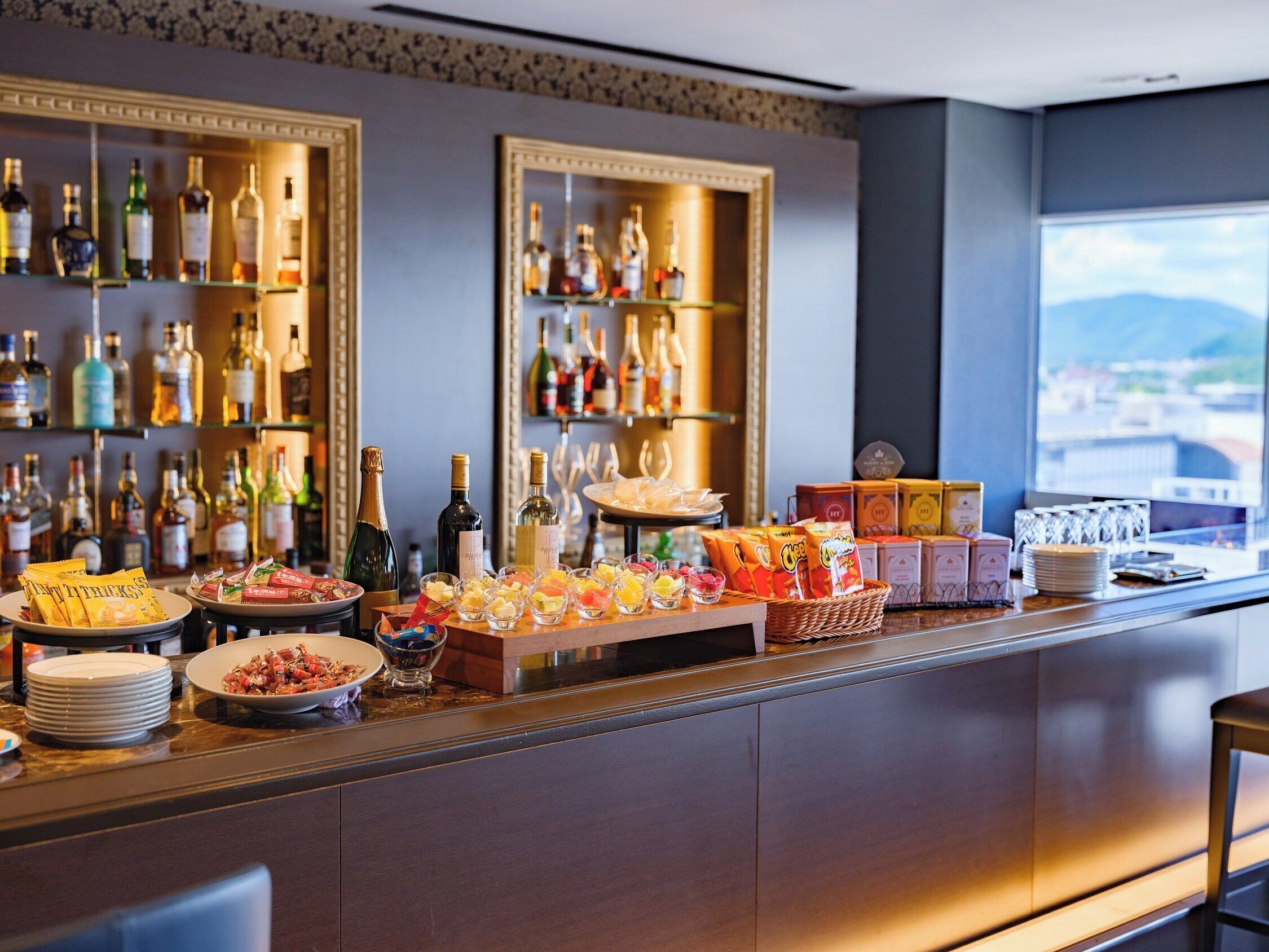 A bar area with shelves of liquor bottles, a variety of snacks, and a view of the city through a large window.