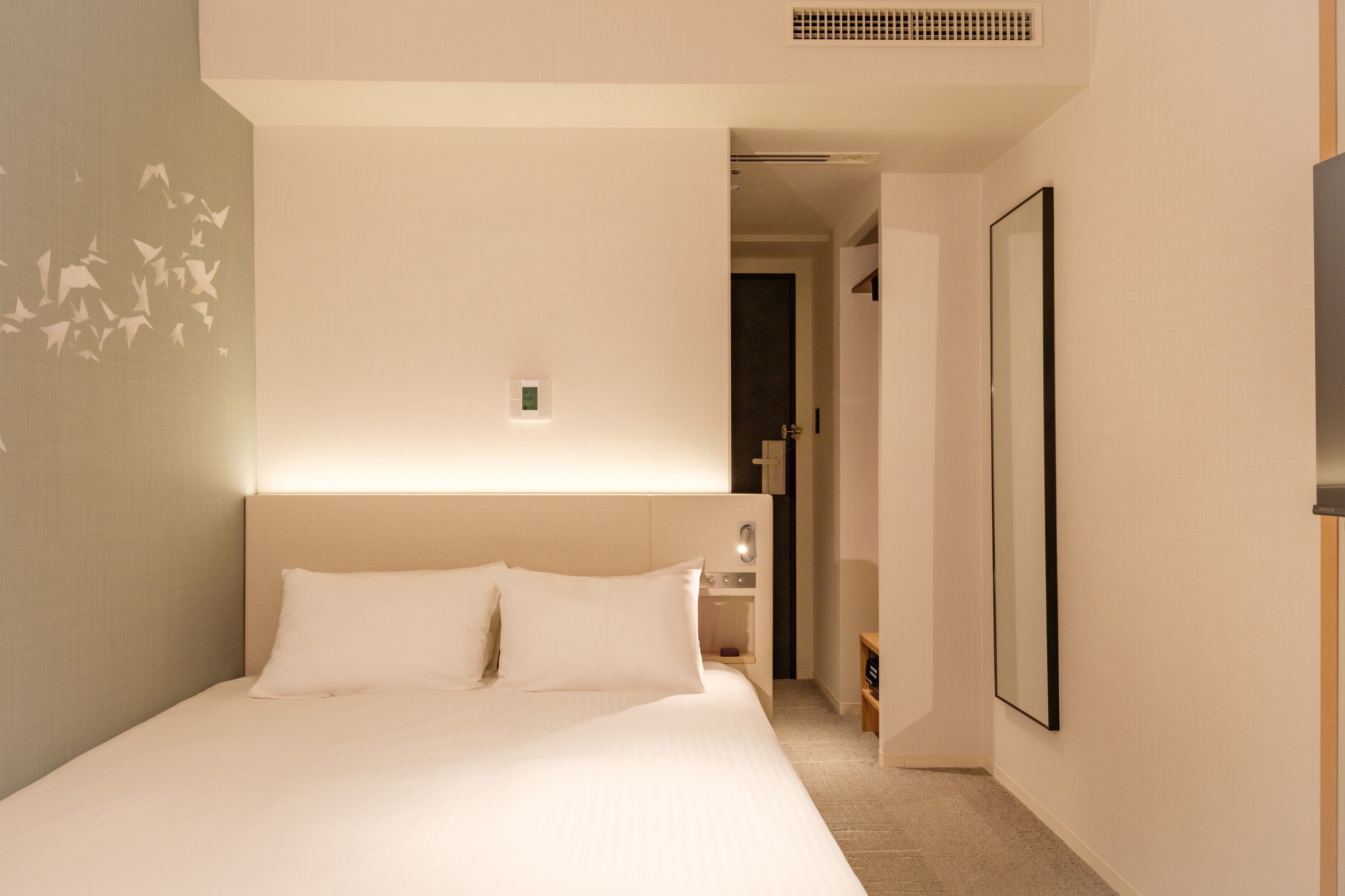 A hotel room with a large bed, a bedside table, a mirror, and a wall-mounted light fixture.