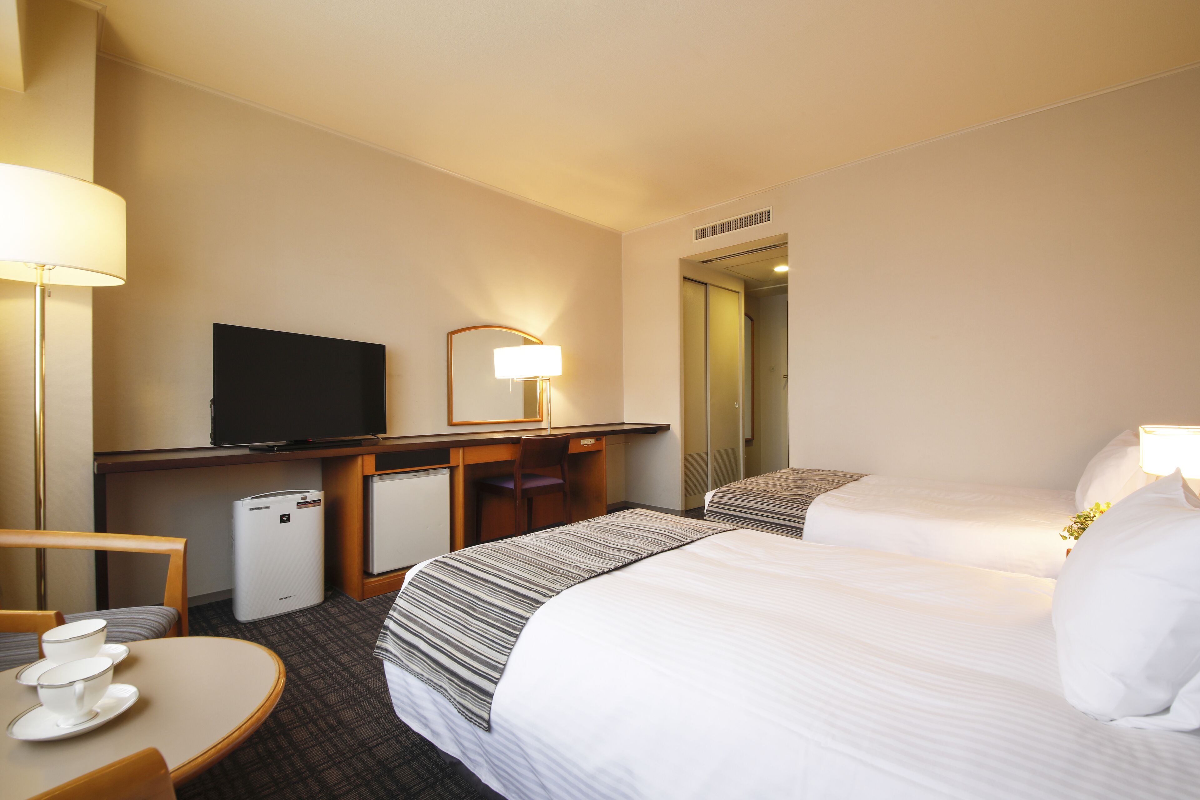 Habitación de hotel con dos camas, un escritorio con televisor, un refrigerador pequeño y una mesa redonda con tazas.