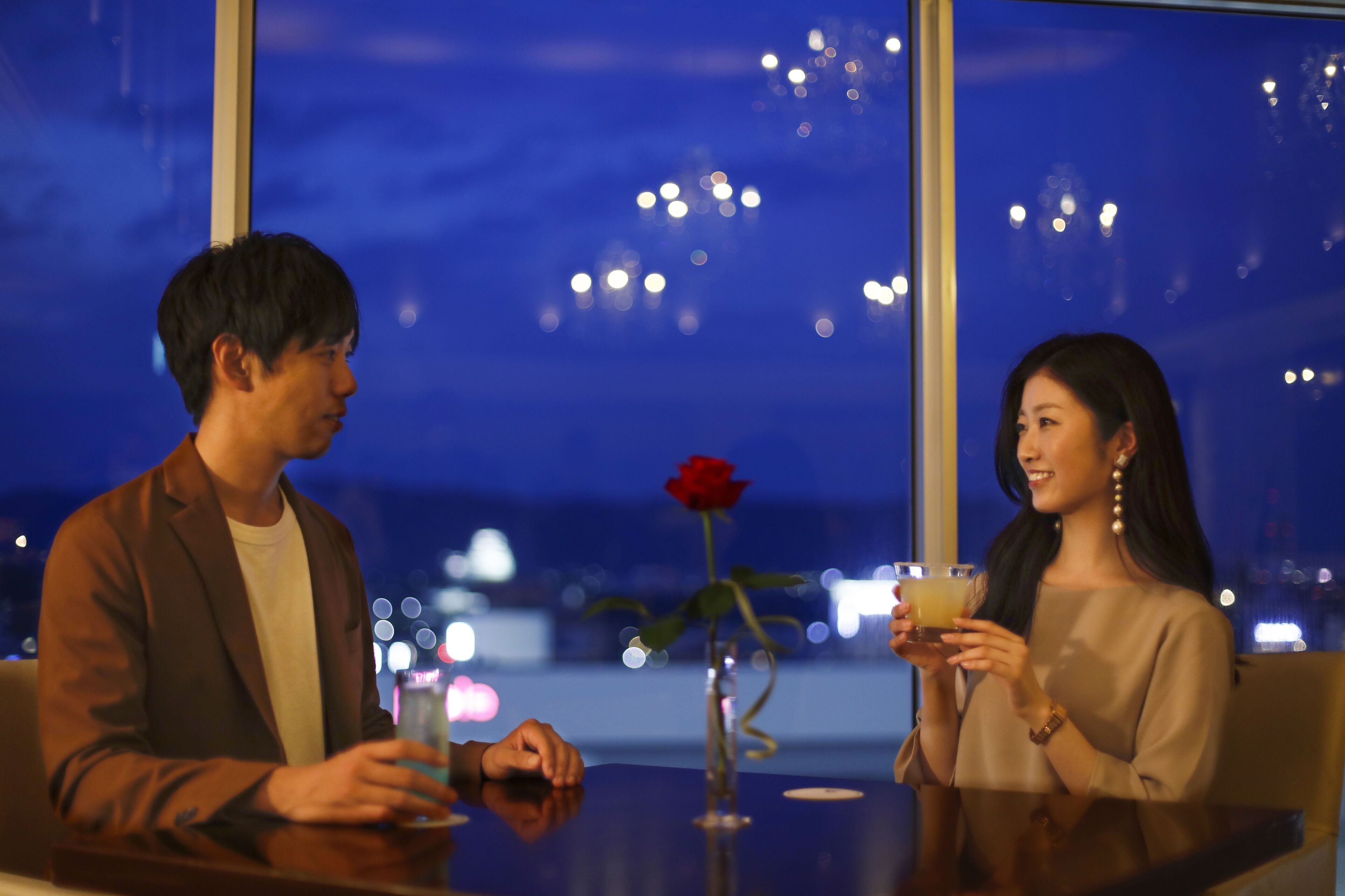 Two people sitting at a table with a vase of a single red rose and a glass of drink.