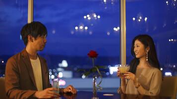 Two people sitting at a table with a vase of a single red rose and a glass of drink.