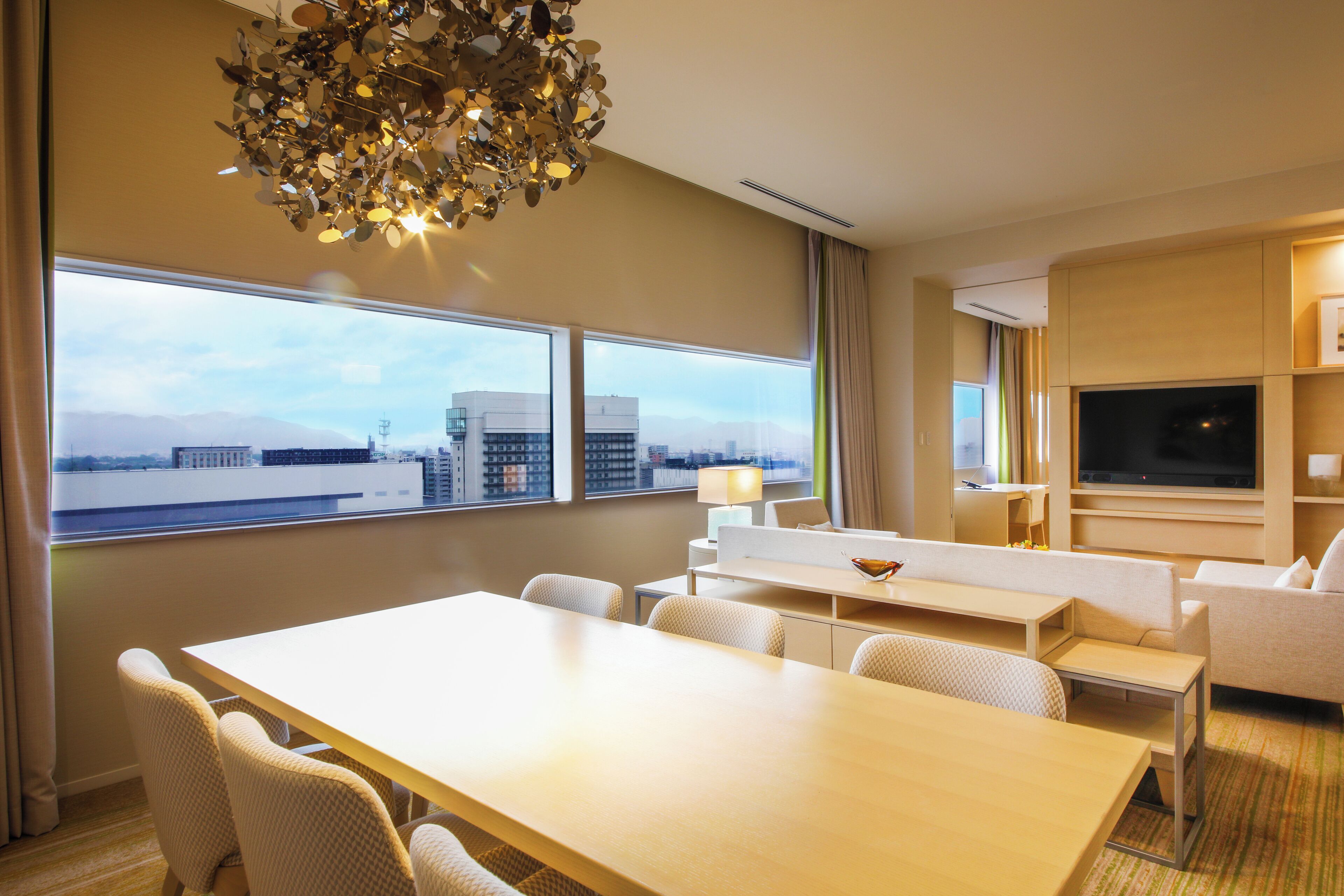 A hotel room with a large window, a dining table, a sofa, a TV, and a chandelier.