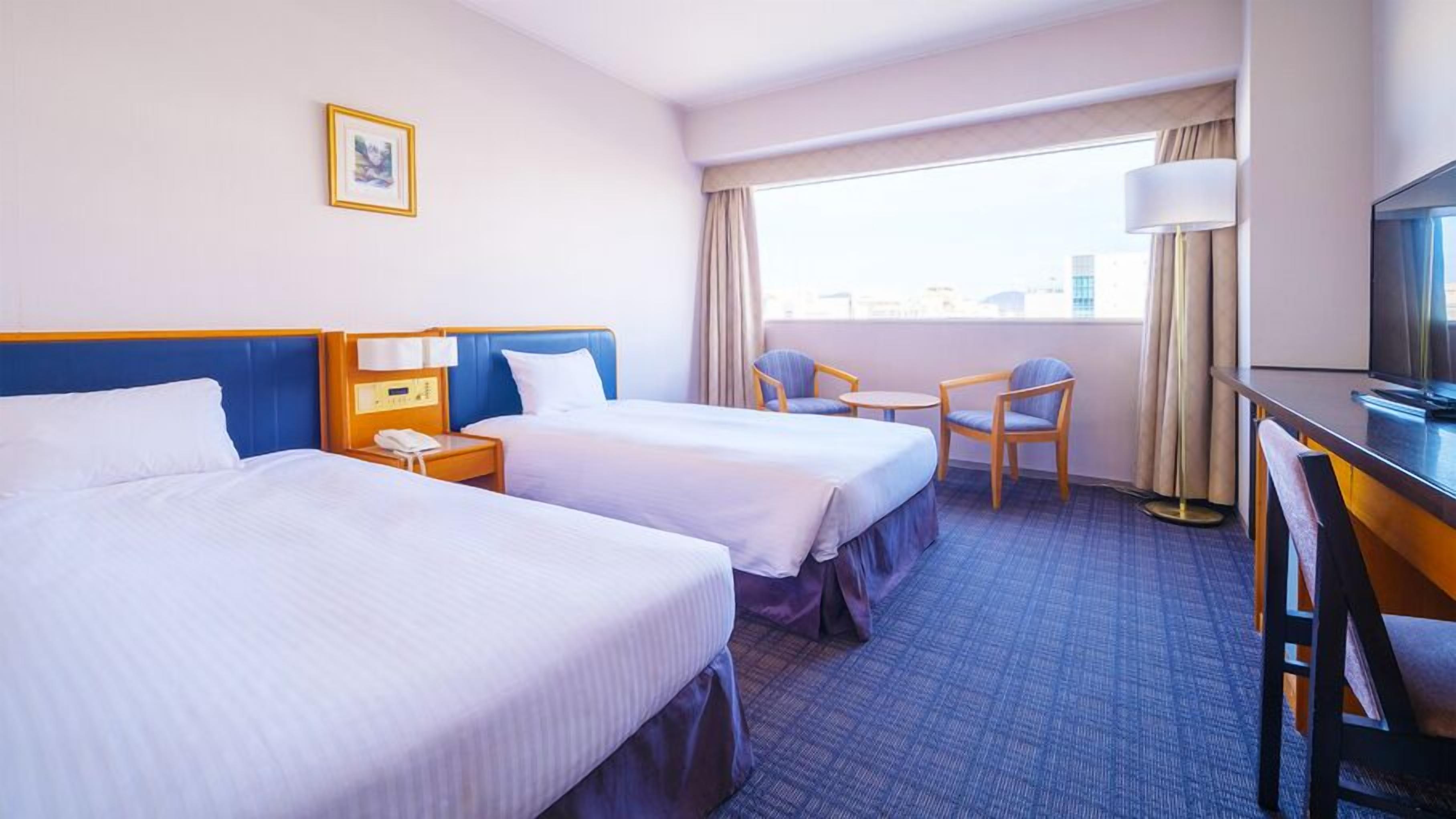 A hotel room with two beds, a desk with a TV, a chair, a small table, and a window with curtains.