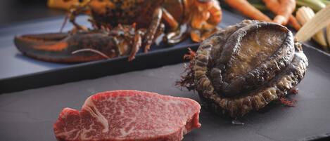 A lobster, a piece of meat, and some vegetables on a dark surface.