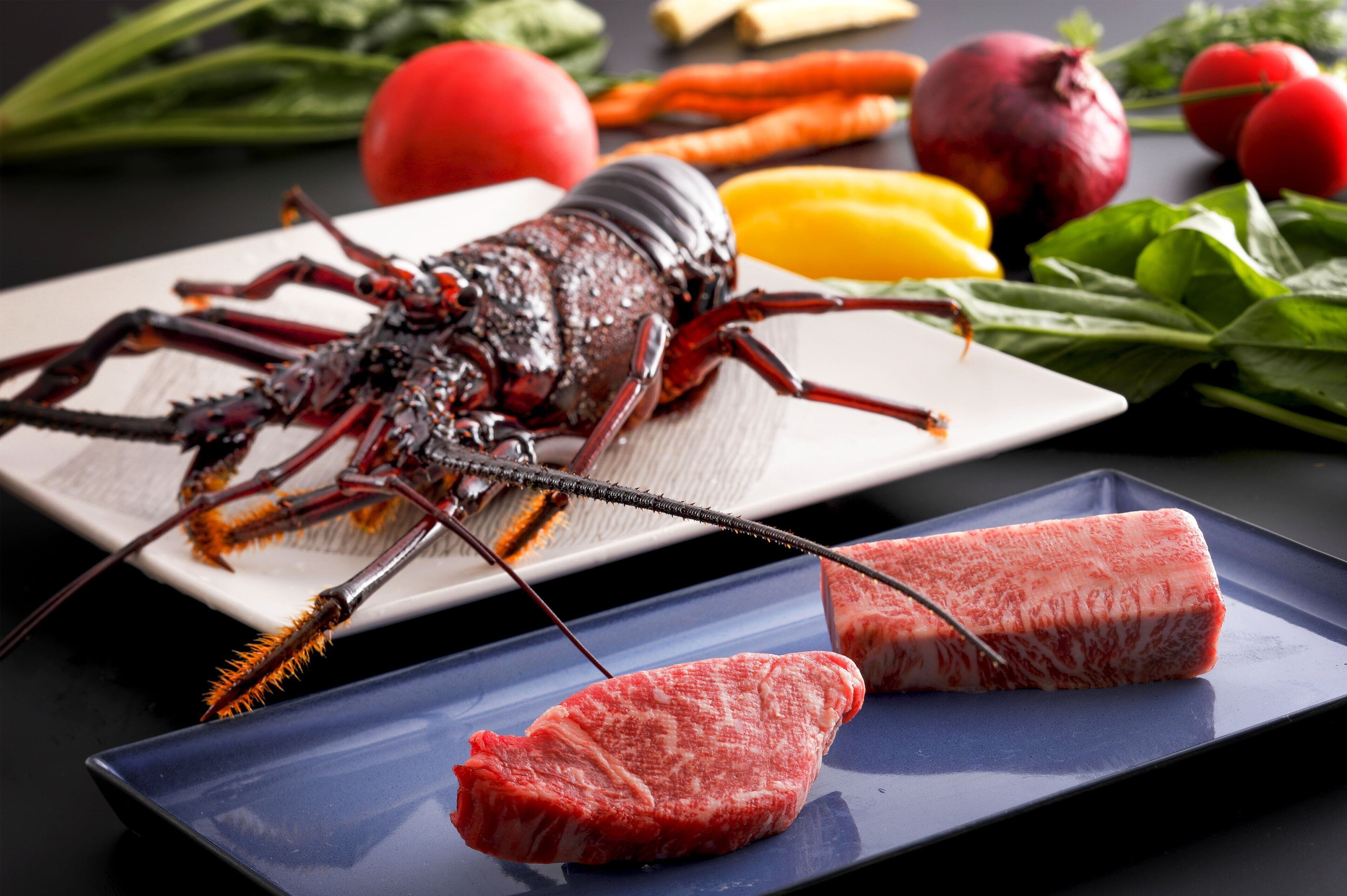 A lobster on a white plate with two slices of red meat on a blue plate, surrounded by vegetables.