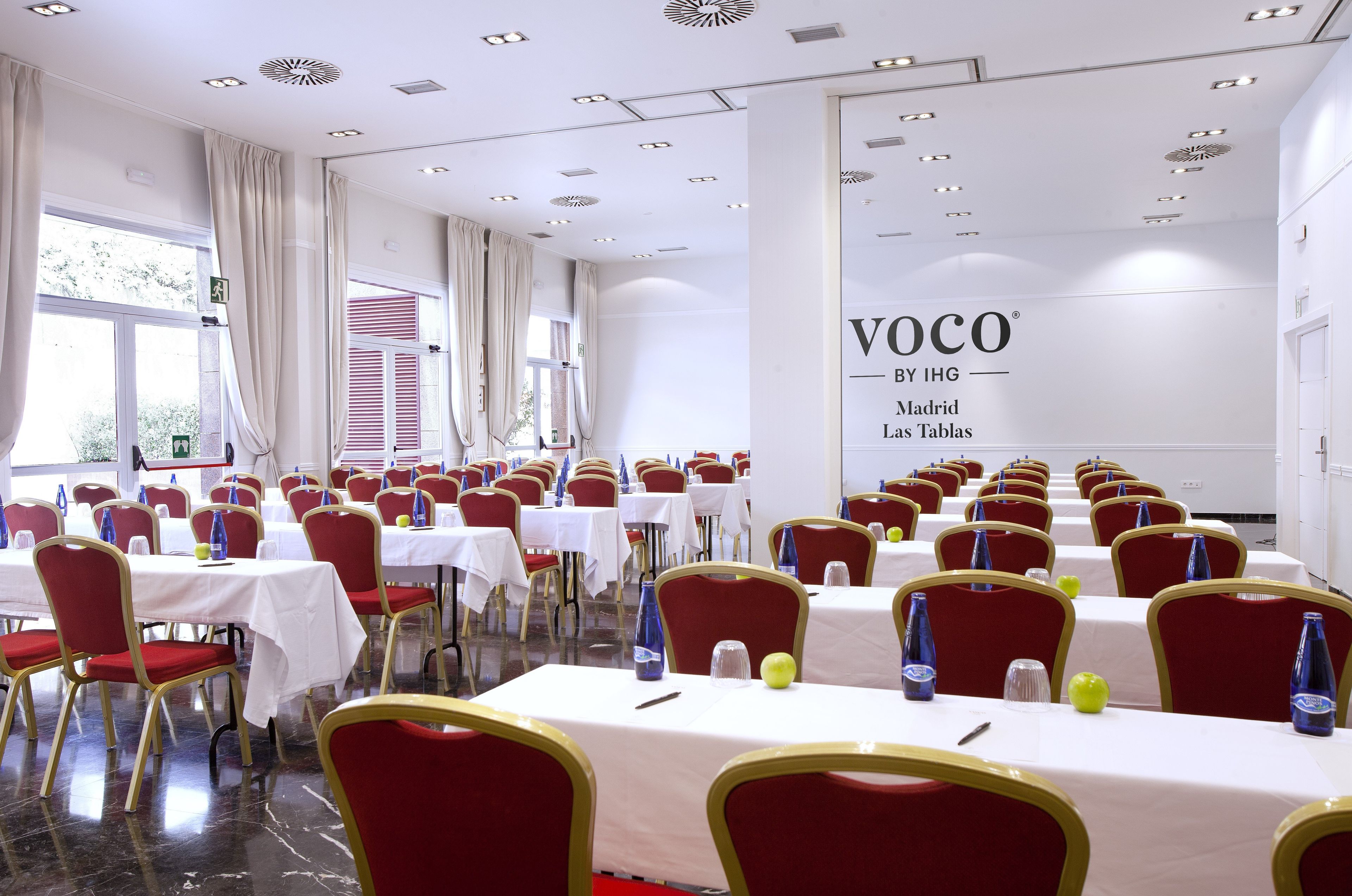 Un salón de conferencias con mesas y sillas, un ventanal con cortinas y una pared con un letrero.