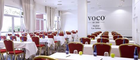 Un salón de conferencias con mesas y sillas, un ventanal con cortinas y una pared con un letrero.
