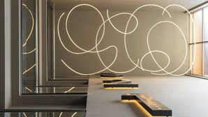 Un interior moderno con diseño minimalista, que presenta un escritorio alargado y estrecho con iluminación integrada, una gran instalación de luces abstracta en la pared y una barandilla de vidrio.