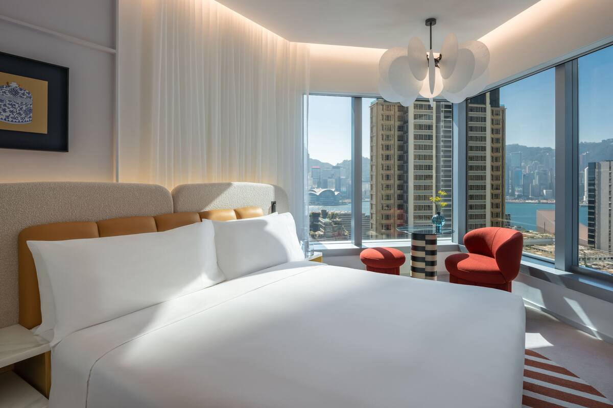 A modern hotel room with a large bed, a red armchair, and a view of tall buildings.