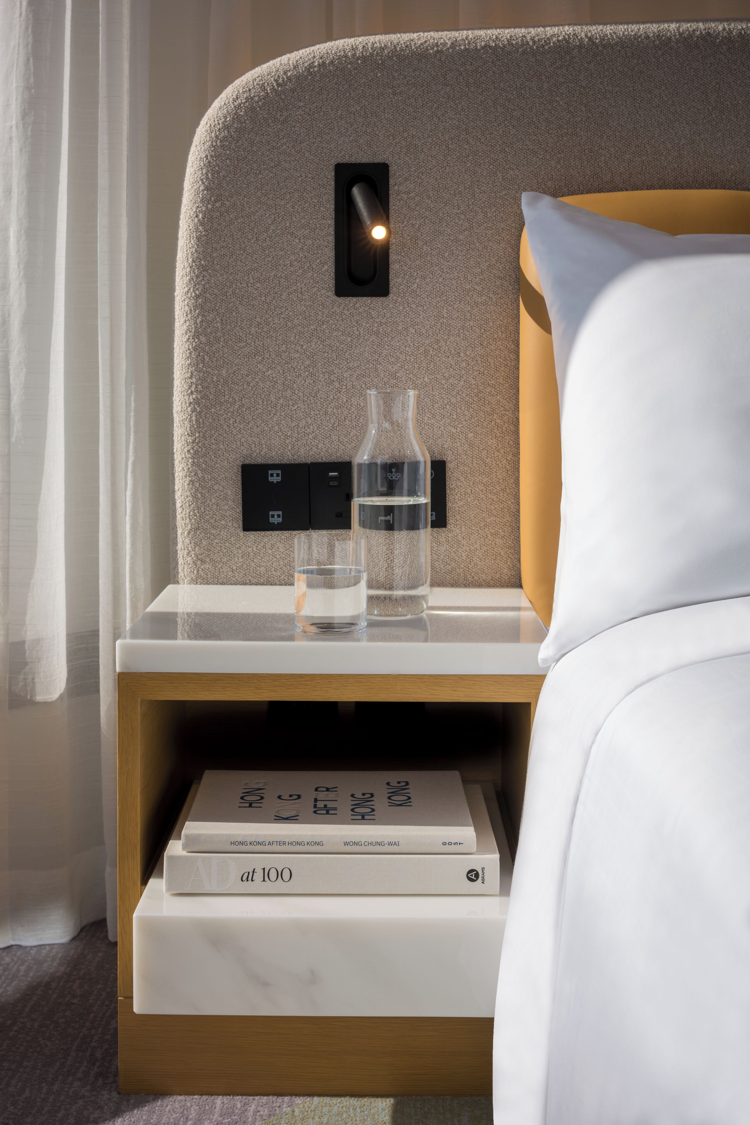 Ein ordentlich bezogenes Hotelbett mit Kopfteil, ein Nachttisch mit einer Wasserflasche, einem Glas und einem Buch mit dem Titel „at 100“. 