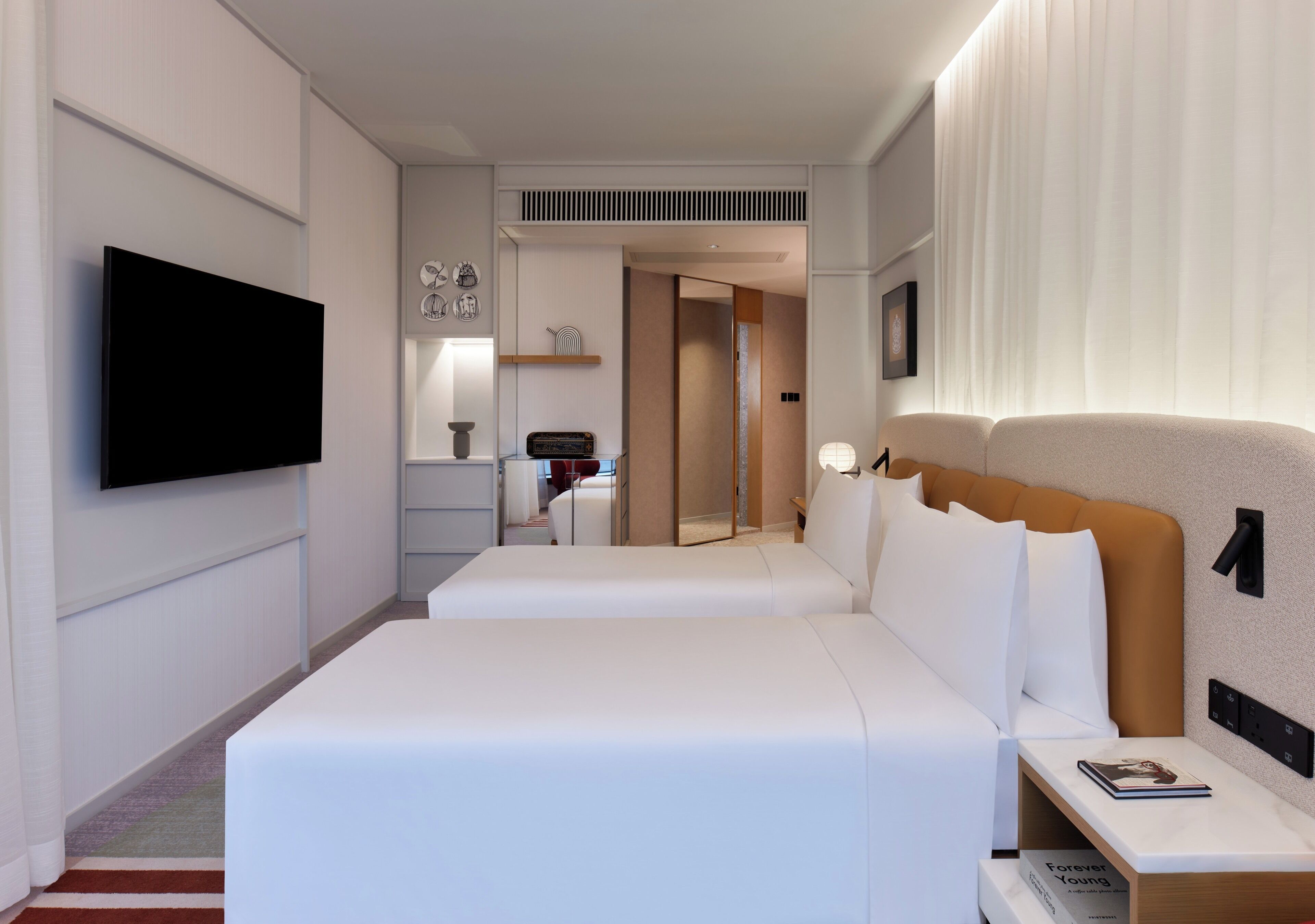 A hotel room with two beds, a flat-screen TV, a bedside table with a book, and a wall-mounted shelf with decorative items.