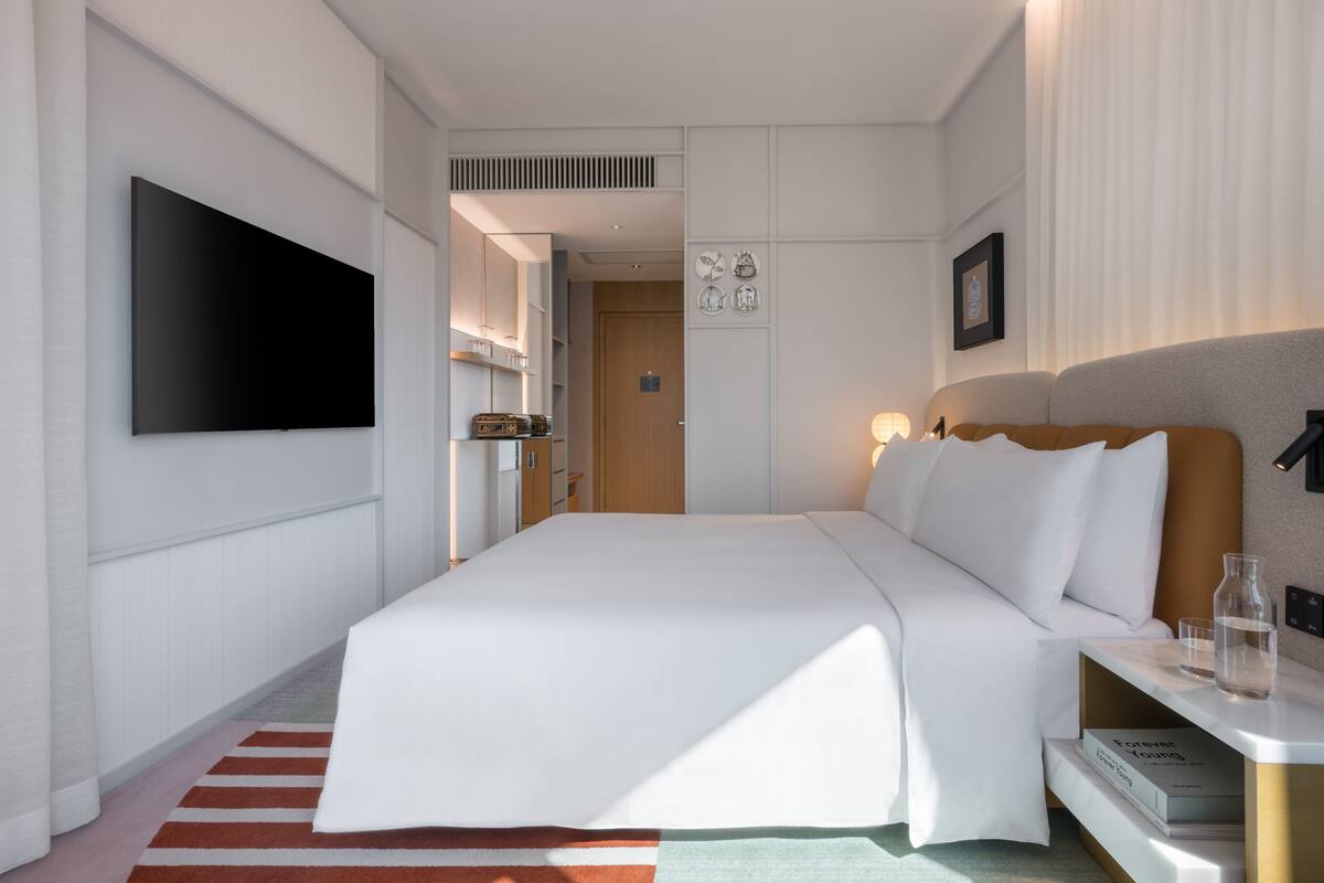 A modern hotel room with a large bed, a flat-screen TV mounted on the wall, a bedside table with a book and a glass, and a door leading to a bathroom.
