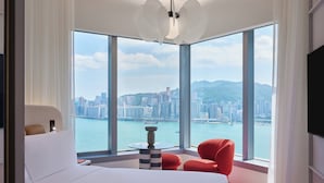 A modern hotel room with a large window offering a city view, a bed, a red chair, and a small table. - Mondrian Hong Kong (Kowloon)