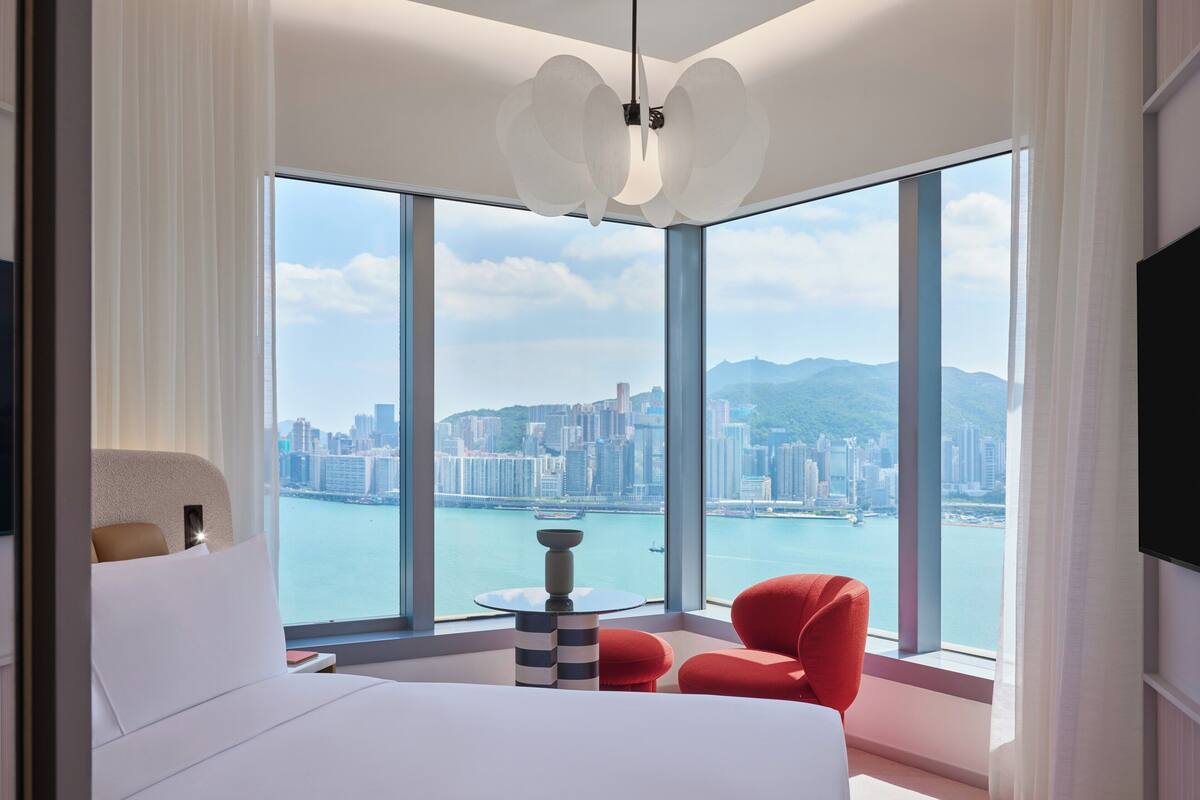 A modern hotel room with a large window offering a city view, a bed, a red chair, and a small table.
