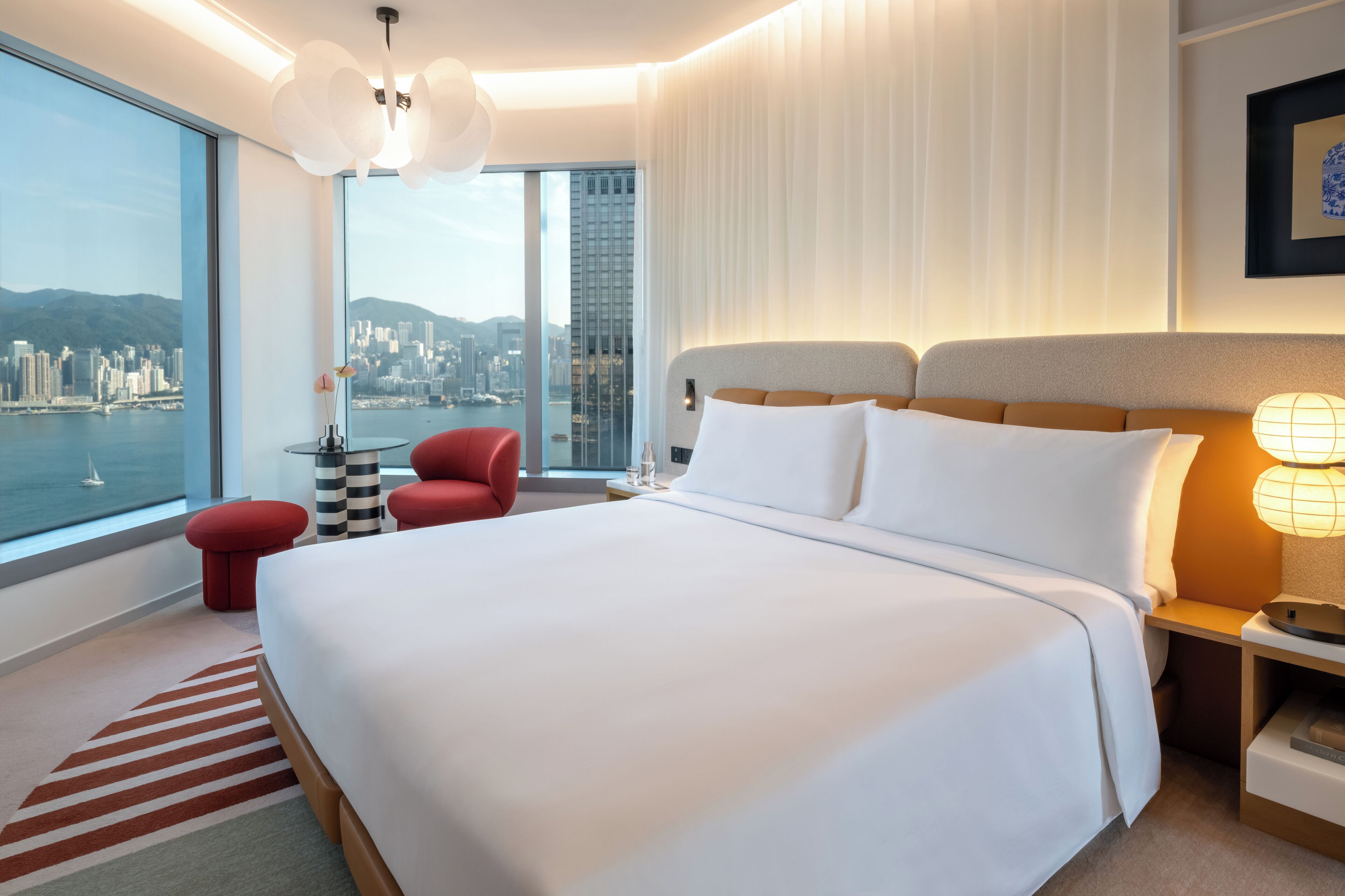 A modern hotel room with a large bed, a red chair, and a view of a cityscape through large windows.