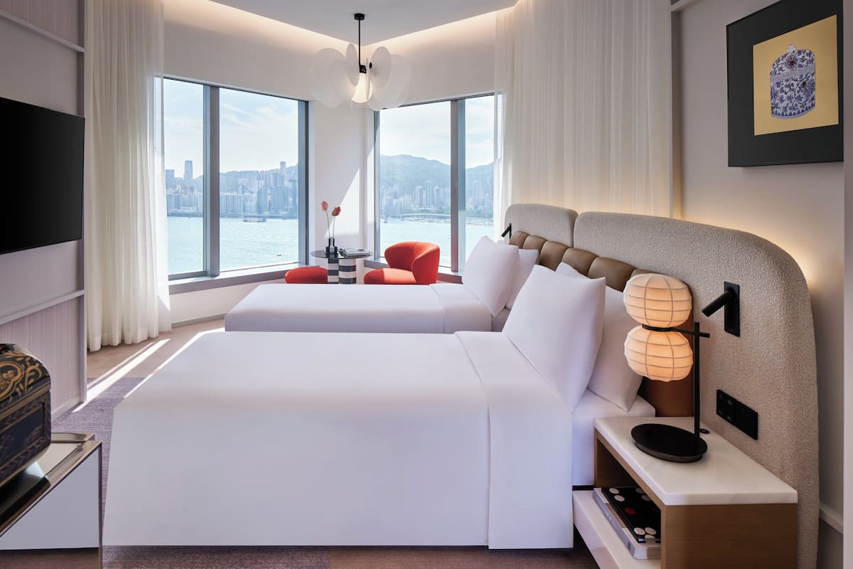 A modern hotel room with a large bed, a seating area with red chairs, a side table with a lamp, and a view of a cityscape through large windows.